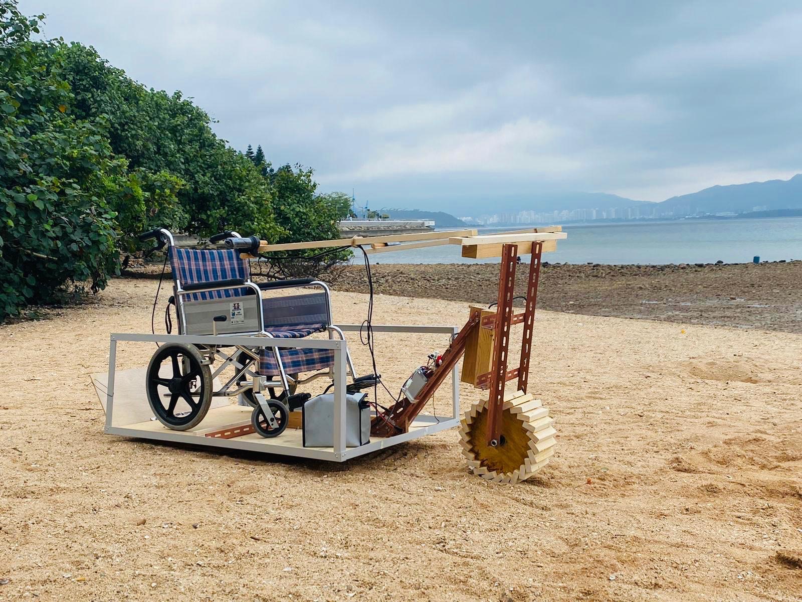Our second-generation vehicle on a beach: a standard wheelchair on a white platform, pushed by a toothed wooden drive wheel.